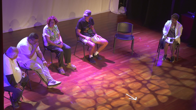 Cinco personas sentadas en sillas sobre un escenario con luz tenue y un ambiente íntimo.